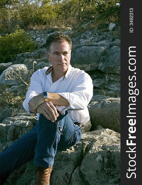 A man, sitting leisurely on a boulder on the side of a rocky hill. A man, sitting leisurely on a boulder on the side of a rocky hill.