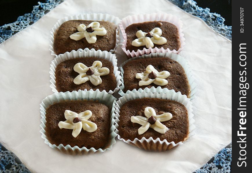 Sitting on a white table cloth with blue lace are six brown cupcakes topped with white frosting flowers with chocolate drops in the center. Sitting on a white table cloth with blue lace are six brown cupcakes topped with white frosting flowers with chocolate drops in the center.