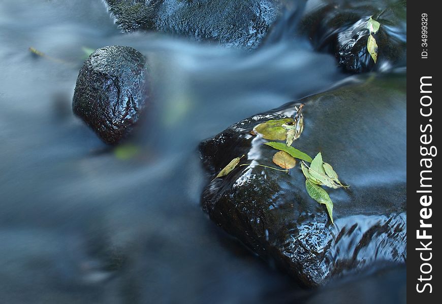 Broken twig on wet stone below increased water level. Blurred motion of blue waves around the stone.