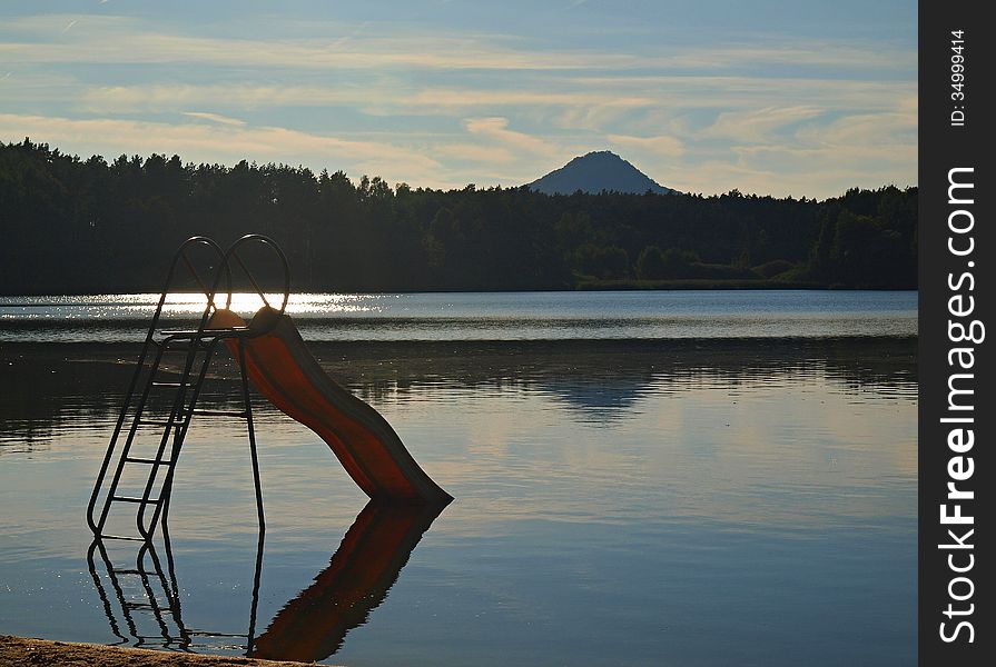 Abandoned Old Children Slide Stays In Pond At The End Of Summer , Reflection In Blue Water Level, Forest At Horizon.