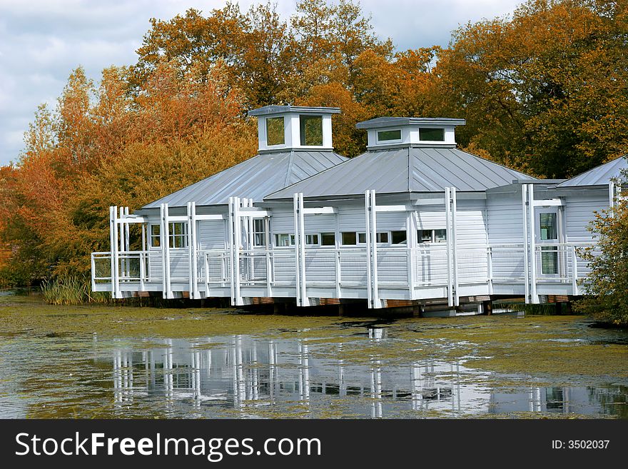 White wooden lakeside residence in autumn with a veranda, with water and pond weed to the foreground and trees to the rear.