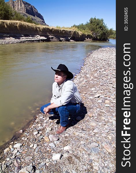 A man dressed in western garb squatting beside a river with an appreciative expression on his face. A man dressed in western garb squatting beside a river with an appreciative expression on his face.