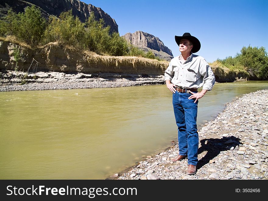 A man dressed in western garb standing beside a river surveying the scene. A man dressed in western garb standing beside a river surveying the scene.