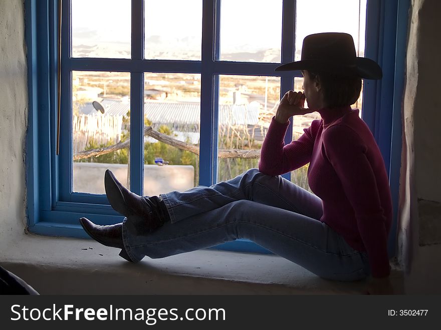 A woman in western apparel sitting on a wide windowsill gazing out the window.