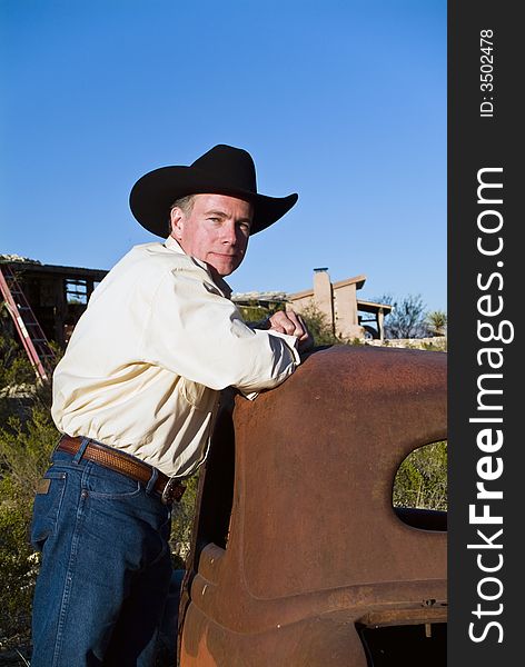 A friendly looking man in a black cowboy hat leaning on an old, long abandoned car. A friendly looking man in a black cowboy hat leaning on an old, long abandoned car.