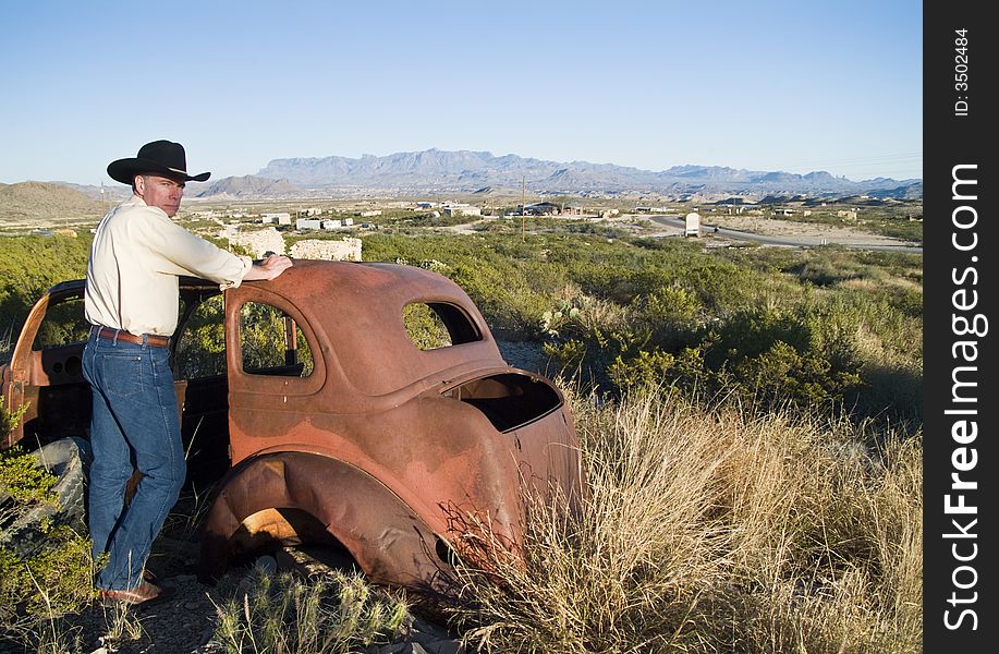 A man in western attire leaning on an old, long abandoned vehicle surrounded by rugged terrain.