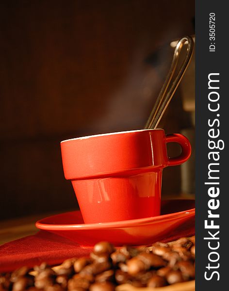 Warm atmosphere for a close-up of a red cup of coffee. Out of focus a moka on the background and coffee beans in first plan to complete the composition. Warm atmosphere for a close-up of a red cup of coffee. Out of focus a moka on the background and coffee beans in first plan to complete the composition.