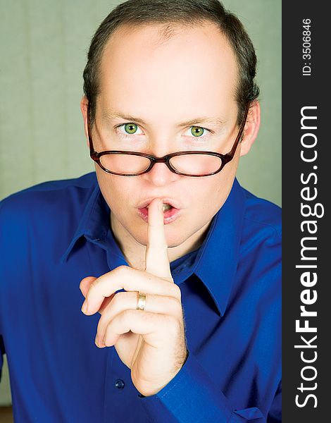 Man with green eyes in blue shirt wearing glasses demanding silence with finger next to his mouth. Man with green eyes in blue shirt wearing glasses demanding silence with finger next to his mouth