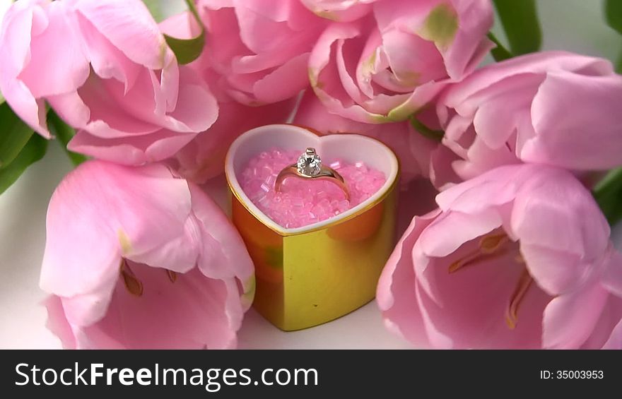 A golden ring with precious stones in gift box as a heart, surrounded by fresh and delicate flowers tulips rotate. A golden ring with precious stones in gift box as a heart, surrounded by fresh and delicate flowers tulips rotate