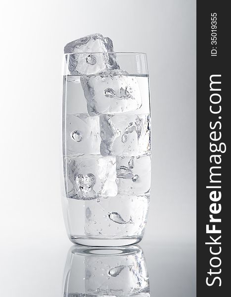 Glass filled with water and ice cubes on a white/grey background, reflection at the bottom. Glass filled with water and ice cubes on a white/grey background, reflection at the bottom