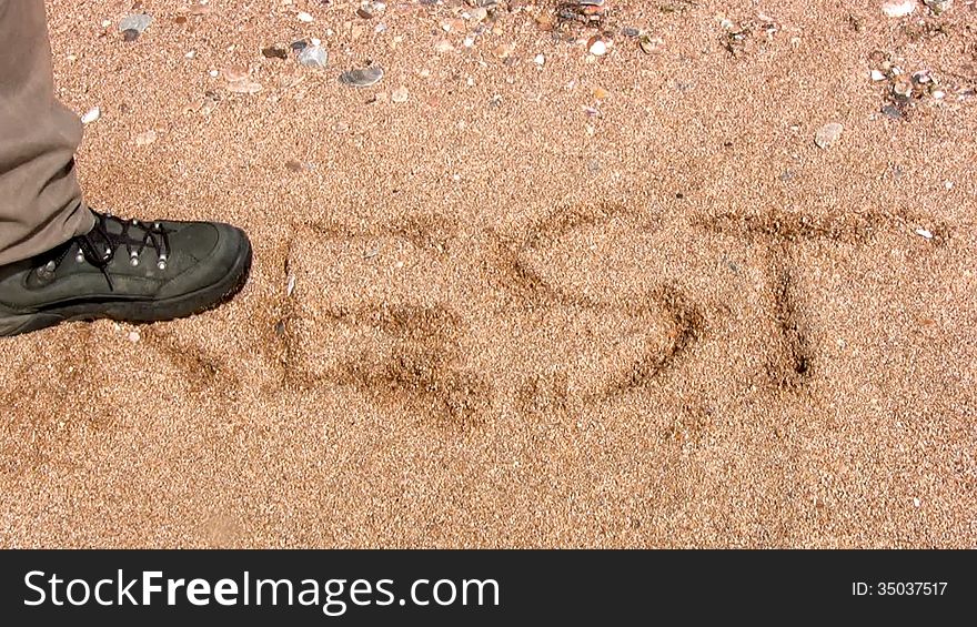 Men's feet in the heavy working boots trample the word rest, written on a yellow sand beach. Men's feet in the heavy working boots trample the word rest, written on a yellow sand beach