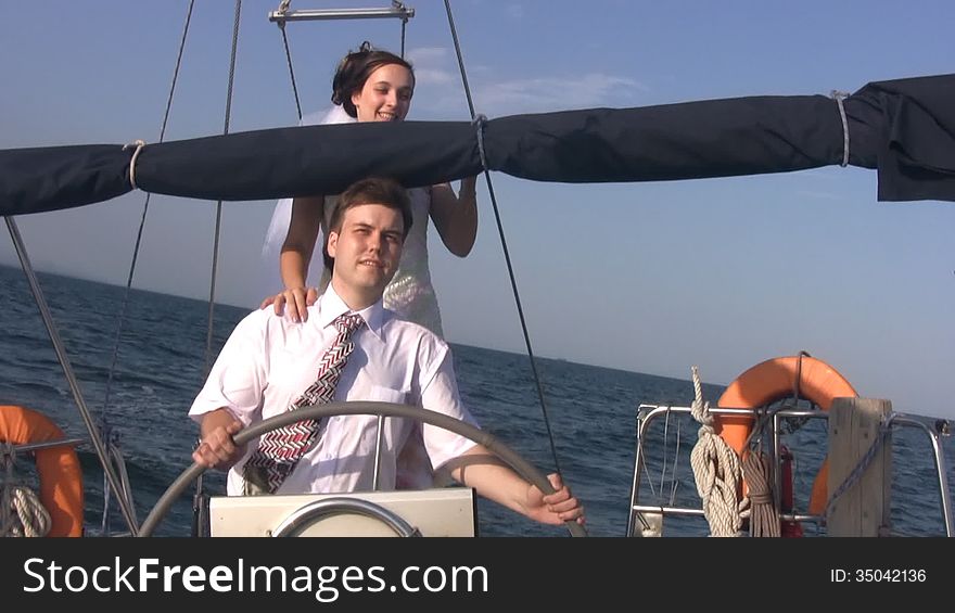 Young happy groom (in white shirt and tie) and young happy bride (in white dress and bridal veil) behind the steering wheel yacht, traveling by sea waves on the background of blue sky. Sun shines. Young happy groom (in white shirt and tie) and young happy bride (in white dress and bridal veil) behind the steering wheel yacht, traveling by sea waves on the background of blue sky. Sun shines