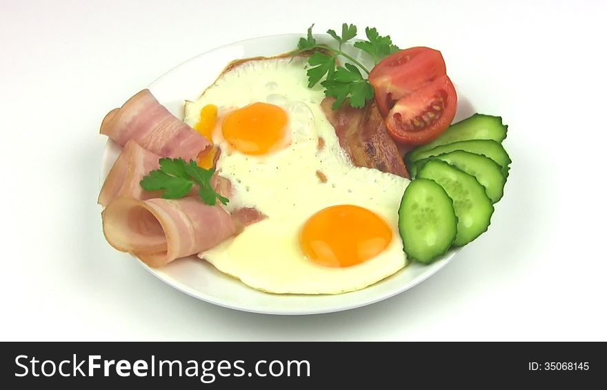 White plate with two fried eggs, bacon, sliced tomato and cucumber, parsley leaves rotates on a white background. Seamless. White plate with two fried eggs, bacon, sliced tomato and cucumber, parsley leaves rotates on a white background. Seamless