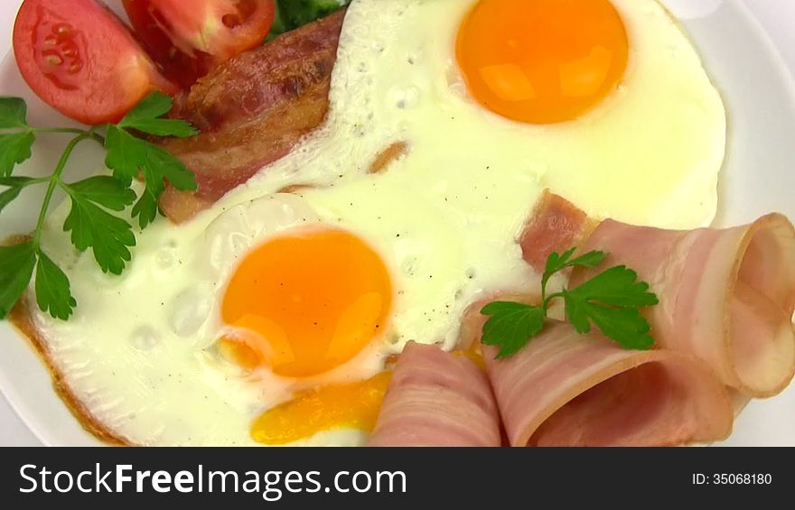 White plate with two fried eggs, bacon, sliced tomato and cucumber, parsley leaves rotates on a white background. Close-up. White plate with two fried eggs, bacon, sliced tomato and cucumber, parsley leaves rotates on a white background. Close-up