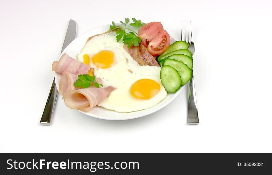 On the white plate is: delicious fried eggs, delicate slices of ham, slices of cucumber and tomato, a sprig of parsley. Near the plates are fork and knife. Plate makes one revolution around its axis