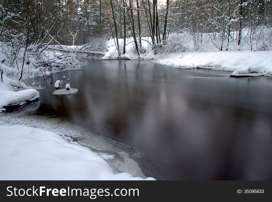 The photograph shows a small river flowing through the forest. It's winter, the ground cover thick layer of snow. The photograph shows a small river flowing through the forest. It's winter, the ground cover thick layer of snow.