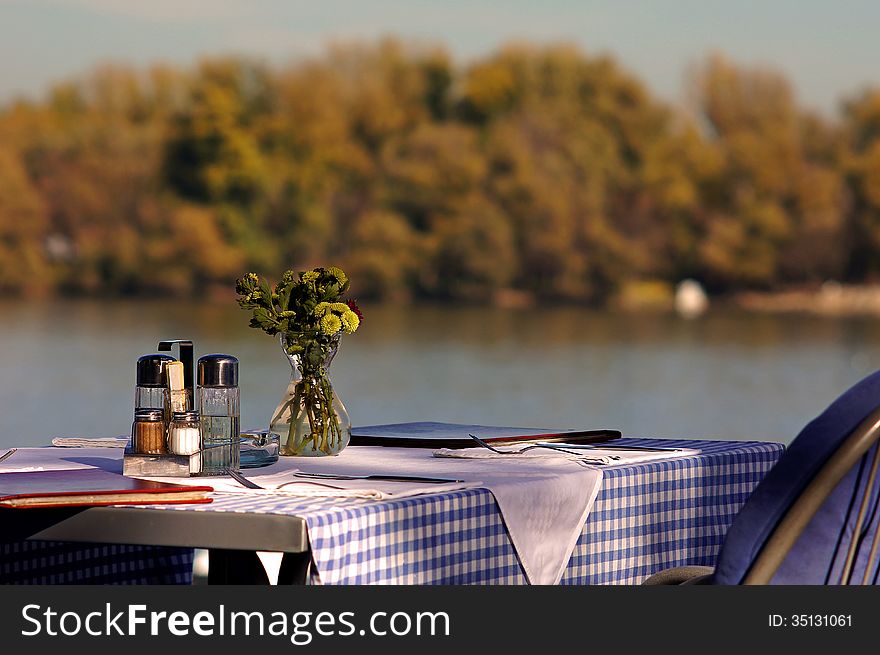 A table at the restaurant at the river bank, set for two, with yellow flowers. A table at the restaurant at the river bank, set for two, with yellow flowers