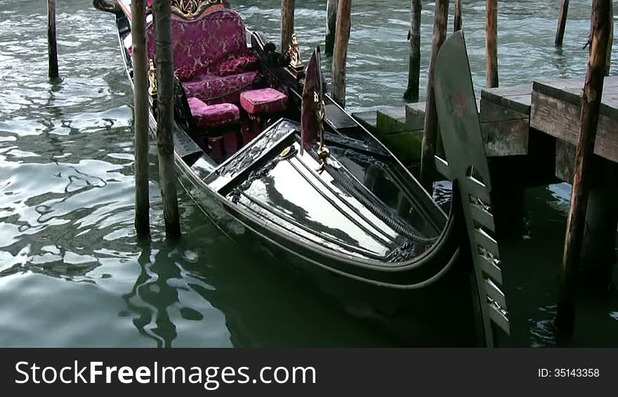 Venice. Grand Canal. Black lacquered gondola sways with the waves. Old wooden pier. Inside the gondola - a red velvet sofa and the flag of Venice. Venice. Grand Canal. Black lacquered gondola sways with the waves. Old wooden pier. Inside the gondola - a red velvet sofa and the flag of Venice