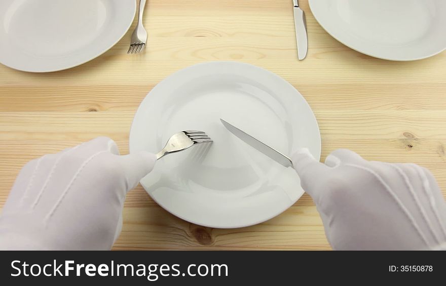 Wooden table. White plate. Hands in white gloves with a fork and a knife cut a piece of invisible food. Wooden table. White plate. Hands in white gloves with a fork and a knife cut a piece of invisible food