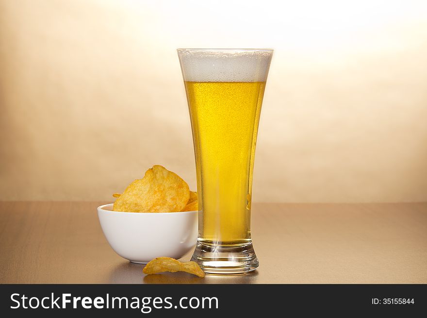 Glass of light amber beer and bowl with chips against brown wall