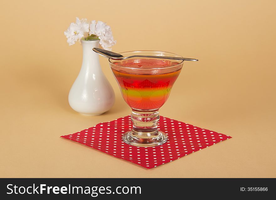 Glass with jelly and a spoon on a napkin, a vase with the flowers, on a beige background. Glass with jelly and a spoon on a napkin, a vase with the flowers, on a beige background