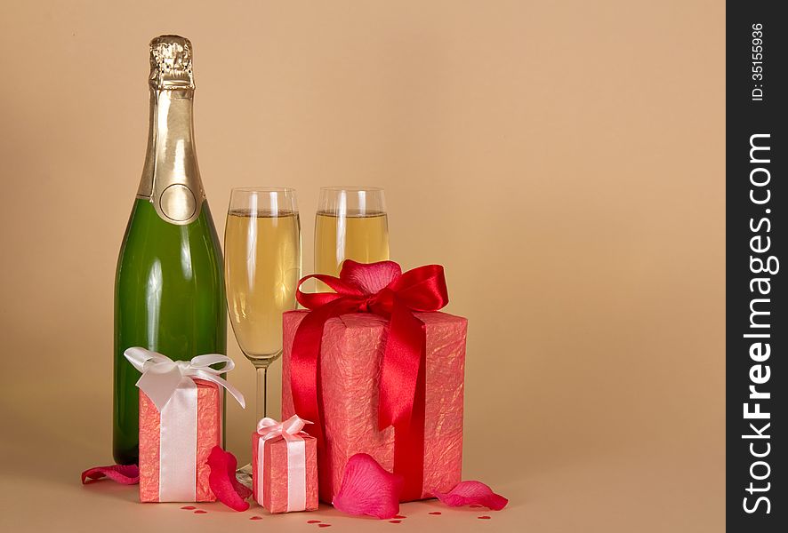 Bottle and wine glasses with champagne, three gift boxes with a ribbon and a bow, rose petals on a beige background