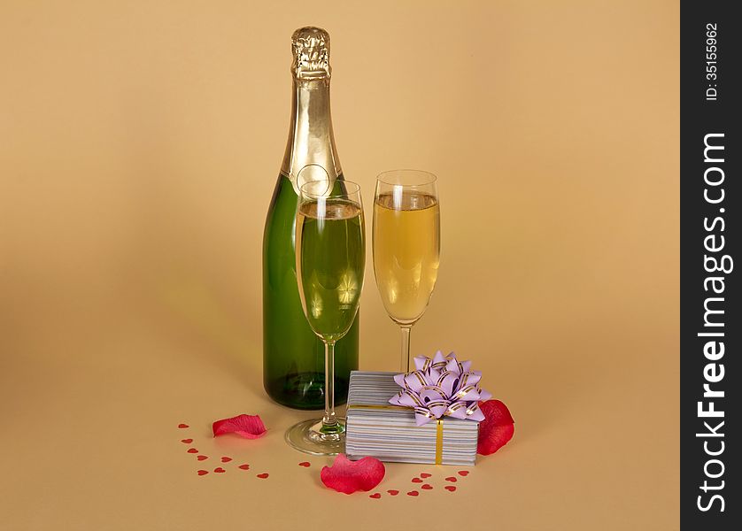 Bottle and two wine glasses of champagne a charming gift box, rose petals and small hearts on a beige background