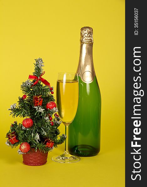 Bottle and wine glass with champagne, and a small fir-tree in a pot on a yellow background