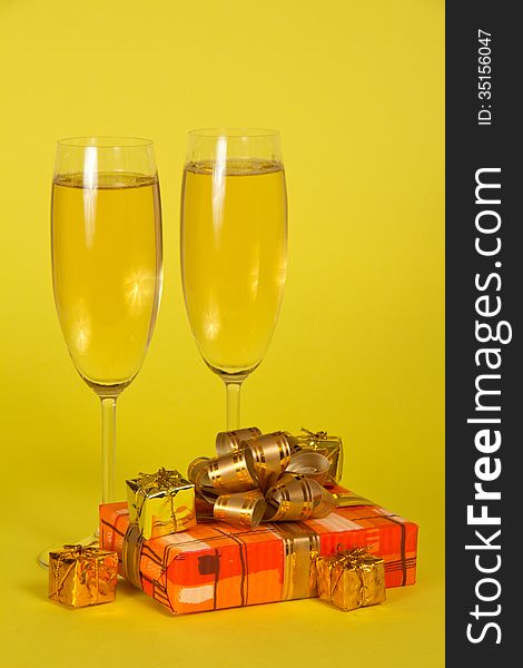 Wine glasses with champagne, a gift box with a beautiful bow and souvenirs on a yellow background. Wine glasses with champagne, a gift box with a beautiful bow and souvenirs on a yellow background