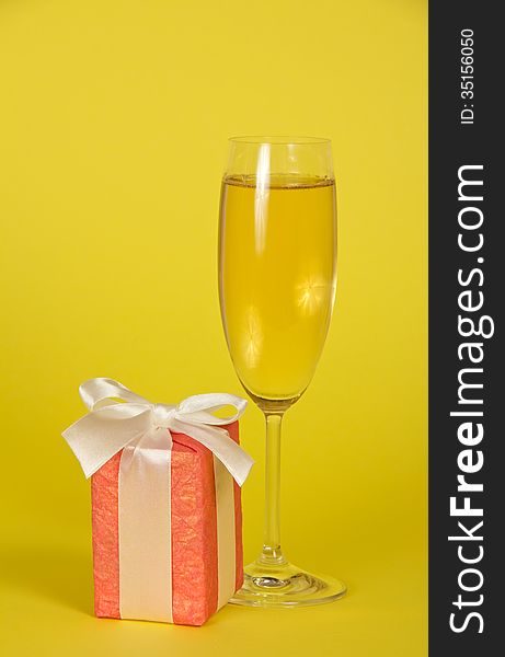 Wine glass with champagne, a gift box with a ribbon and a bow on a yellow background. Wine glass with champagne, a gift box with a ribbon and a bow on a yellow background