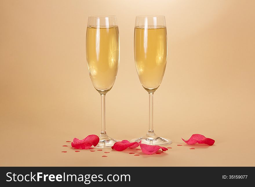 Two wine glasses of champagne, rose petals and small hearts on a beige background. Two wine glasses of champagne, rose petals and small hearts on a beige background