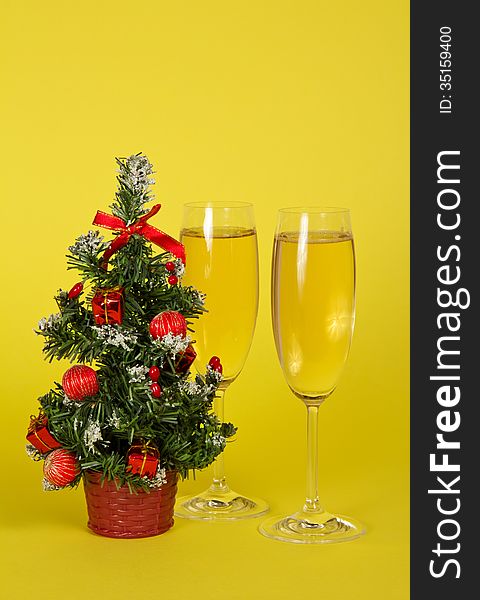 Small Christmas fir-tree in a pot, and two wine glasses with champagne on a yellow background