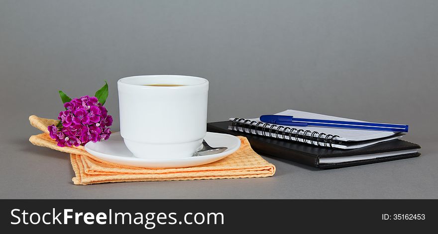 Cup of coffee with a saucer, a spoon and a flower on a napkin, a sketchpad and the ball-point on a gray background. Cup of coffee with a saucer, a spoon and a flower on a napkin, a sketchpad and the ball-point on a gray background