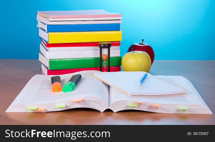 The open book with bookmarks, exercise book a pen and markers, a pile of the closed books a hourglass and apples on a table