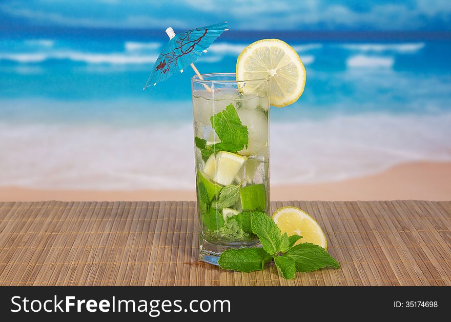 Green cocktail of alcohol with an umbrella, a lemon and spearmint, on a bamboo cloth against the sea. Green cocktail of alcohol with an umbrella, a lemon and spearmint, on a bamboo cloth against the sea