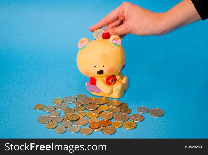 A girl's hand, putting a coin inside a toy safe, with a bunch of coins scattered on a blue background. A girl's hand, putting a coin inside a toy safe, with a bunch of coins scattered on a blue background