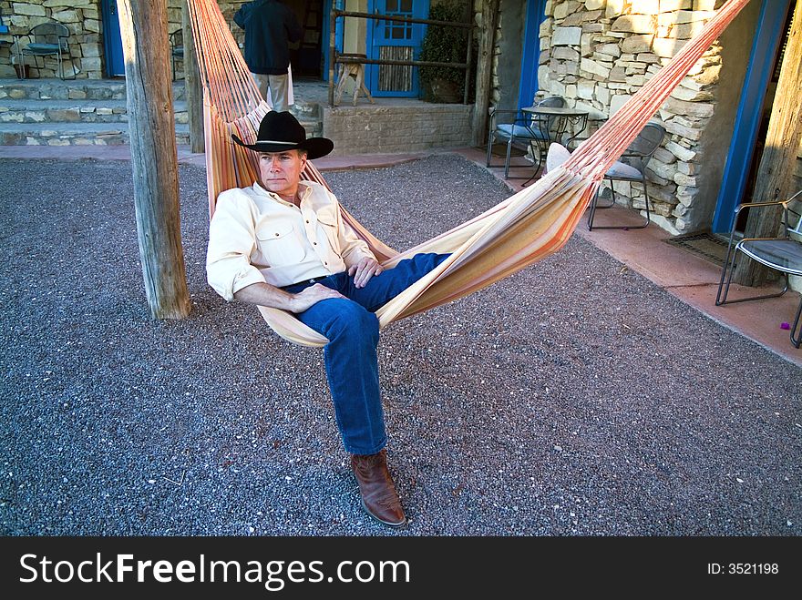 A man dressed in a cowboy hat and boots resting in a colorful hammock. A man dressed in a cowboy hat and boots resting in a colorful hammock.