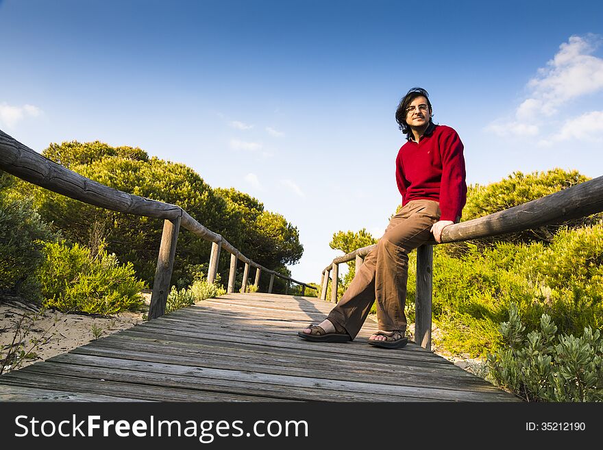 Man sitting on the rustic wooden railings of a coastal boardwalk crossing the dunes and beach sand, as he relaxes and enjoys the sunshine and the tranquillity of nature. Man sitting on the rustic wooden railings of a coastal boardwalk crossing the dunes and beach sand, as he relaxes and enjoys the sunshine and the tranquillity of nature.