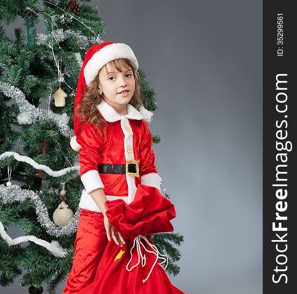 A little girl dressed in a suit Snta Claus standing near a Christmas tree and holding a red bag with gifts. A little girl dressed in a suit Snta Claus standing near a Christmas tree and holding a red bag with gifts