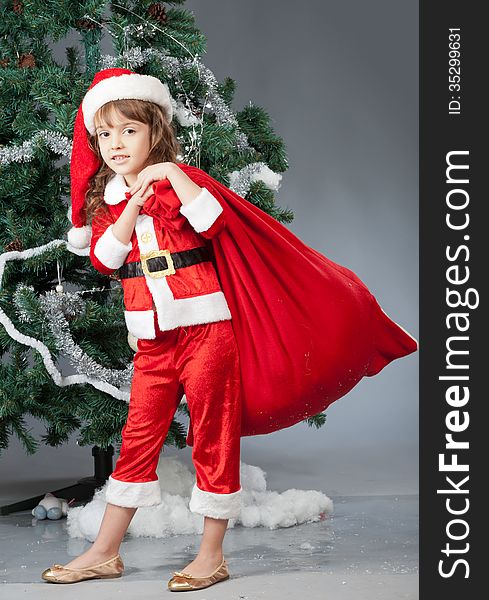 A little girl dressed in a suit Snta Claus standing near a Christmas tree and holding a red bag with gifts. A little girl dressed in a suit Snta Claus standing near a Christmas tree and holding a red bag with gifts