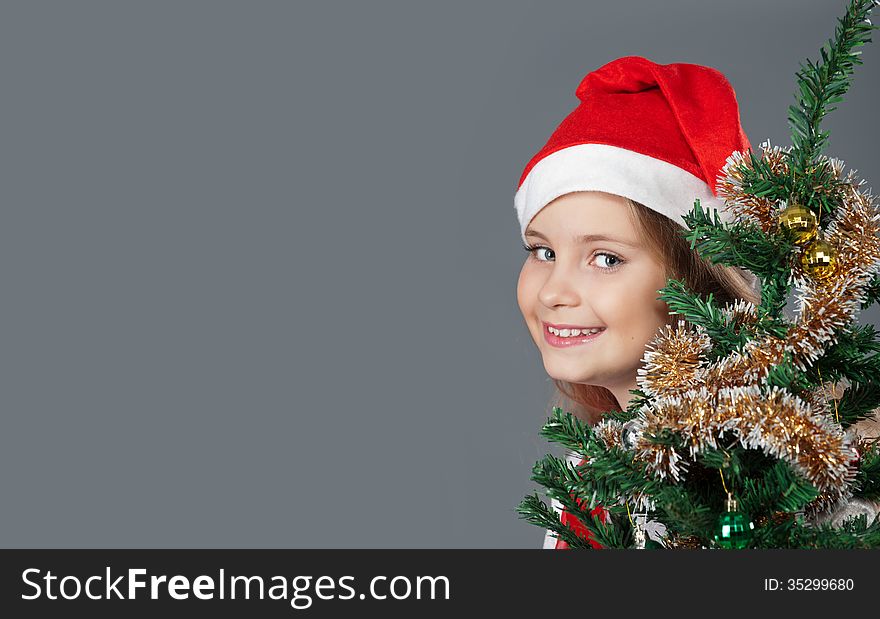 Little girl in Santa hat smiling and peeking out from behind the Christmas tree. Little girl in Santa hat smiling and peeking out from behind the Christmas tree