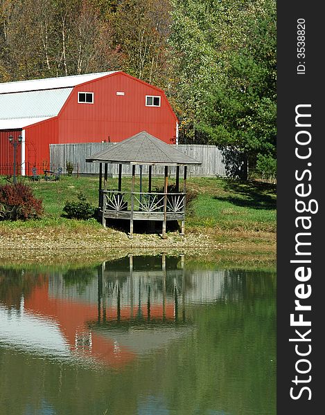 A gazebo on a pond with reflection and a barn behind