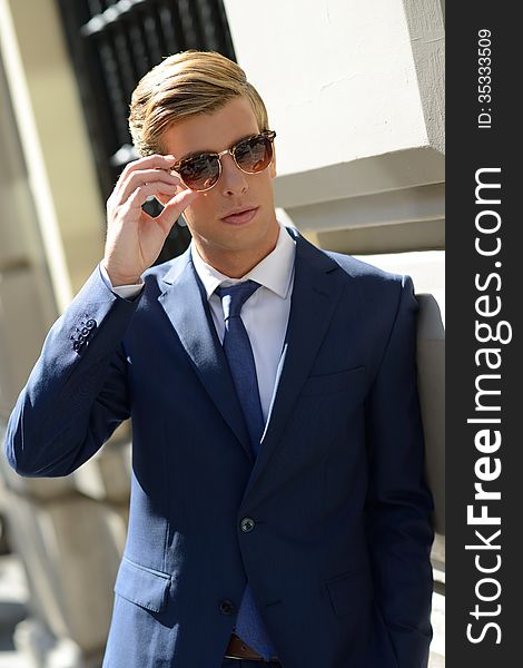 Portrait of an attractive young businessman in urban background wearing blue suit, sunglasses and necktie. Blonde hair