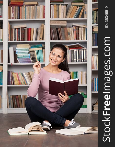 Beautiful young female student holding book and smiling while sitting on the floor at the library. Beautiful young female student holding book and smiling while sitting on the floor at the library