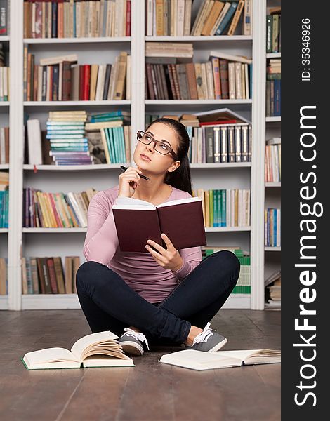 Thoughtful young woman holding a book and looking away while sitting on the floor at the library. Thoughtful young woman holding a book and looking away while sitting on the floor at the library