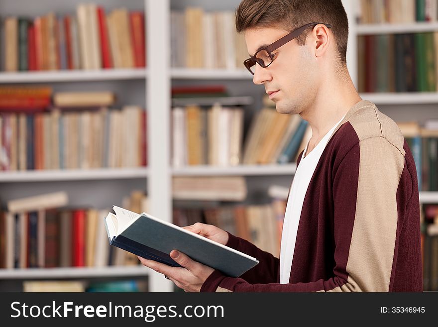 Side View of young man reading a book while standing at the library. Side View of young man reading a book while standing at the library