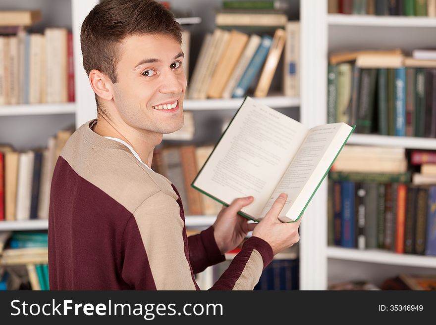 Cheerful young man holding a book and looking over shoulder while standing at the library. Cheerful young man holding a book and looking over shoulder while standing at the library