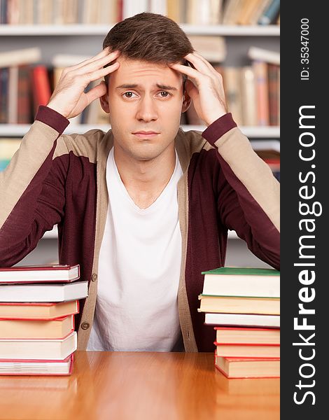 Tired young man holding his head in hands and leaning at the book stack while sitting at the library desk. Tired young man holding his head in hands and leaning at the book stack while sitting at the library desk