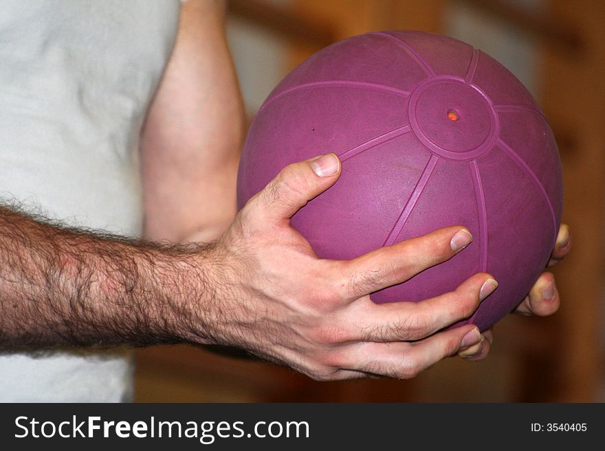 Two hands holding in suspension a purple medical ball for a exercise in the gym / Wall Bars in Blurred Background. Two hands holding in suspension a purple medical ball for a exercise in the gym / Wall Bars in Blurred Background
