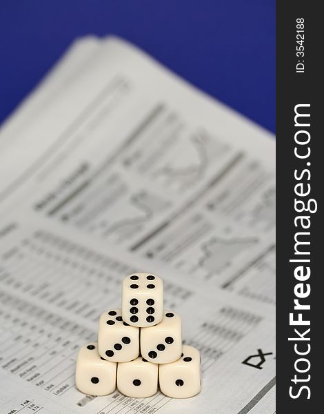 A set of six dice forming a little pyramid with six dots on the top one, lying on newspaper - financial markets section, blue background. A set of six dice forming a little pyramid with six dots on the top one, lying on newspaper - financial markets section, blue background.